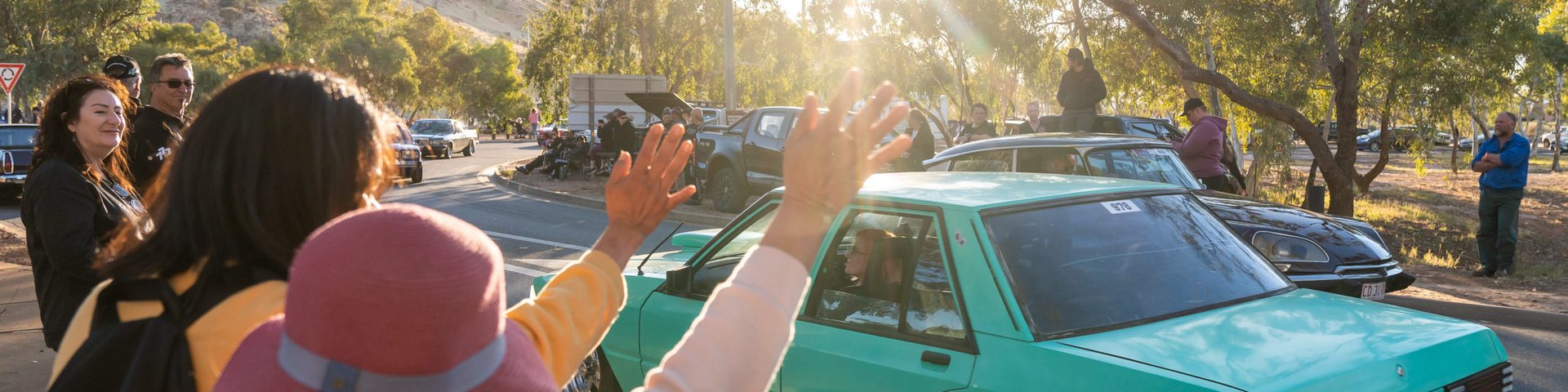 Crowd waving as a teal sedan cruises past during the street parade, spectators lining a tree-shaded road with parked cars behind.