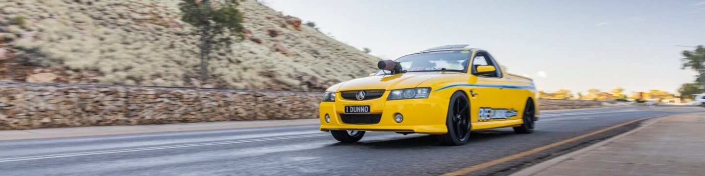 Yellow ute driving an outback highway past red ranges, front plate reading '1 DUNNO'.