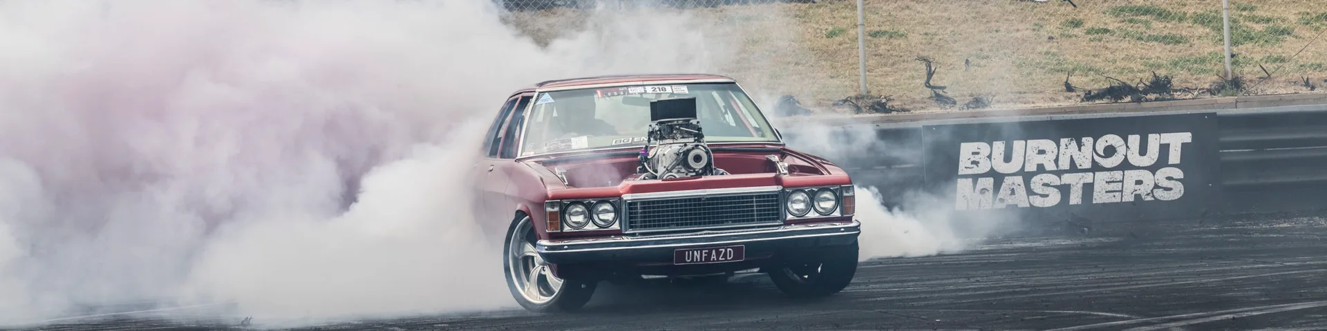 Dark red vintage sedan with a supercharger doing a burnout in smoke, plate 'UNFAZD', beside a 'Burnout Masters' sign.