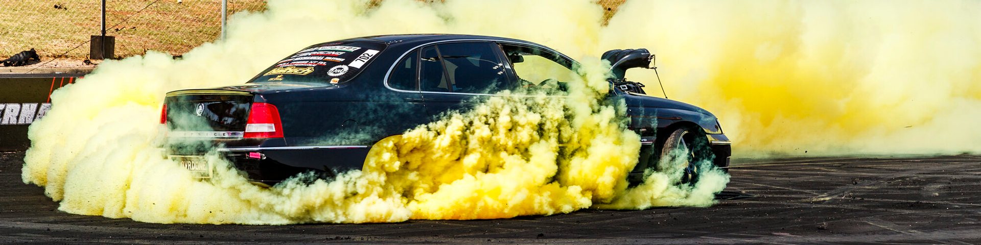 Dark sedan doing a burnout in thick yellow-tinted smoke on a drag strip, a partial sponsor banner visible at left.